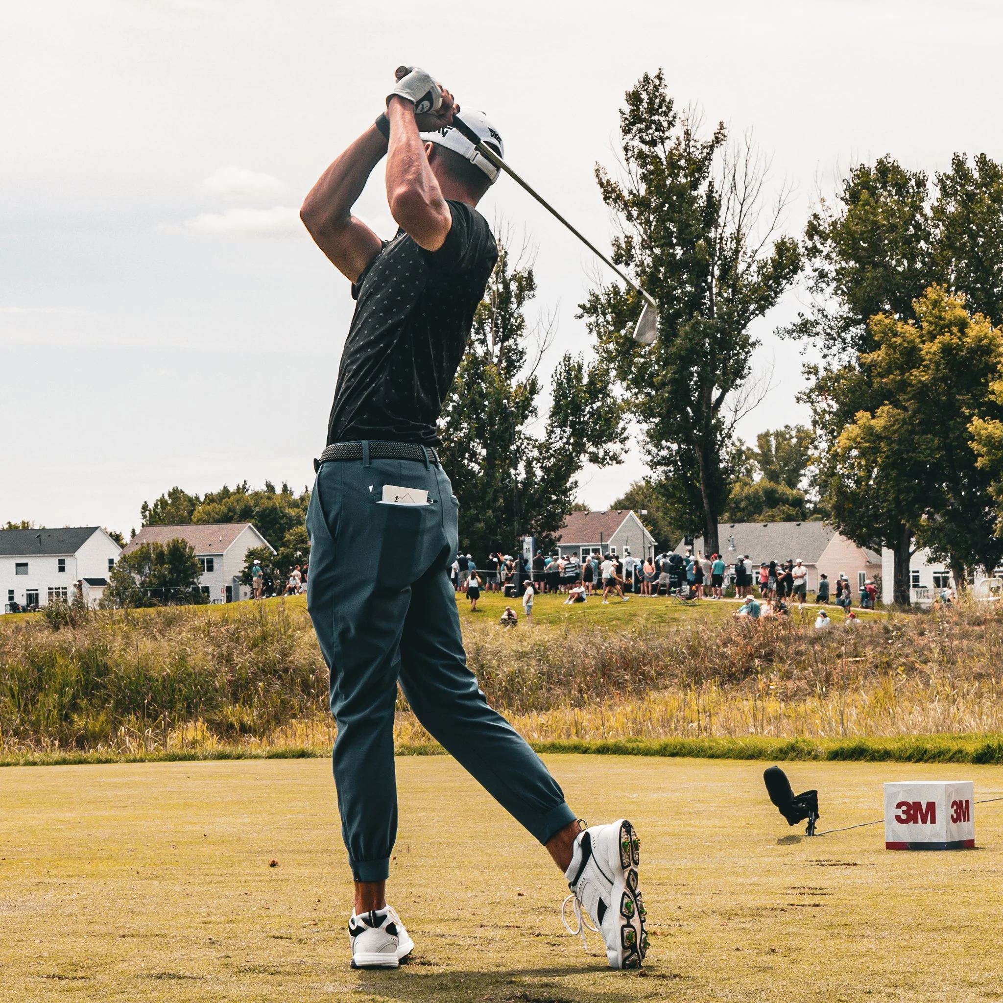 A golfer in a black shirt and blue pants swings a club on a golf course, with a group of people and houses in the background.