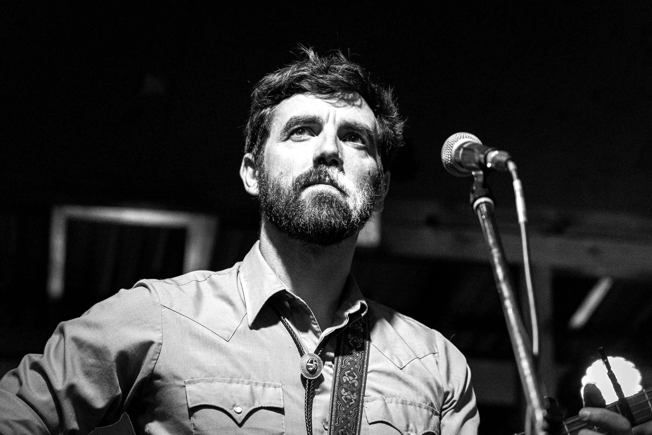 Black and white photo of a man with a beard and mustache singing or performing with a microphone in front of him, holding a guitar, on stage with a dark background.