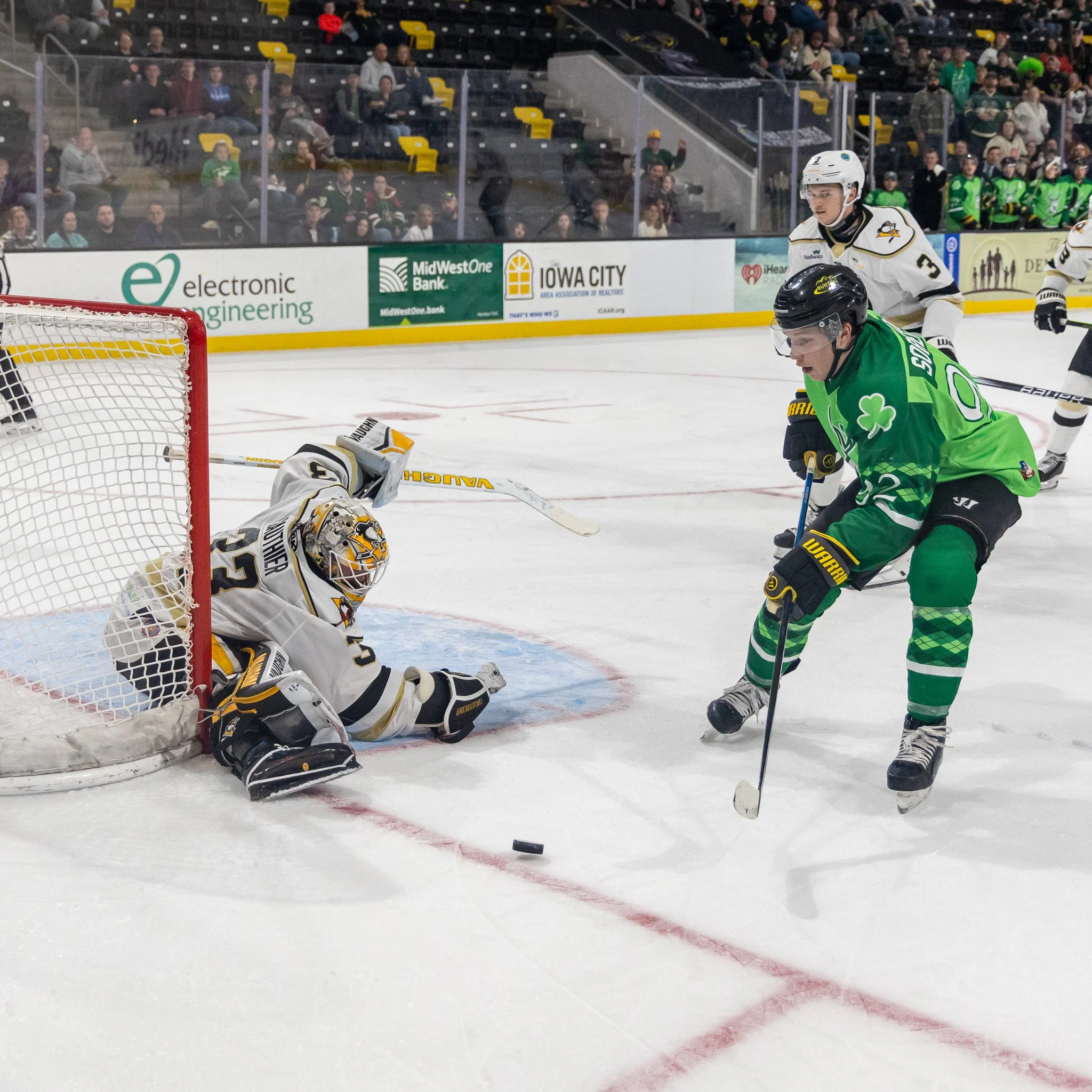 An ice hockey game with a player in green attempting to shoot the puck past the goalie in white, who is on the ice blocking the shot during a game in an indoor arena with fans and advertising boards in the background.