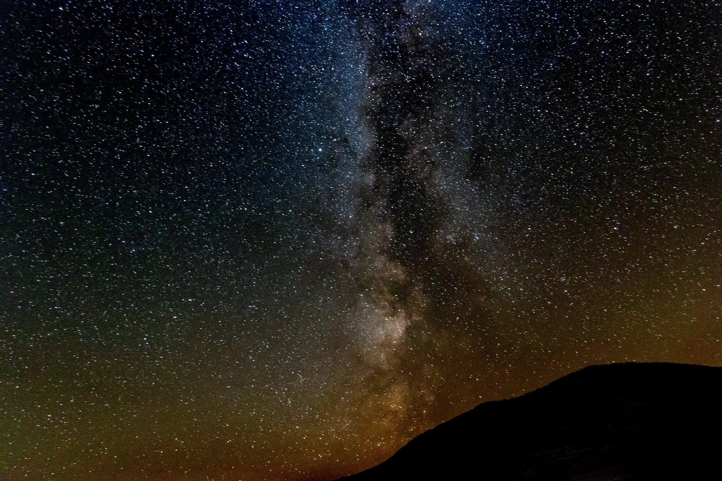 Night sky filled with stars and the Milky Way galaxy, with a silhouette of a hill or mountain in the foreground.