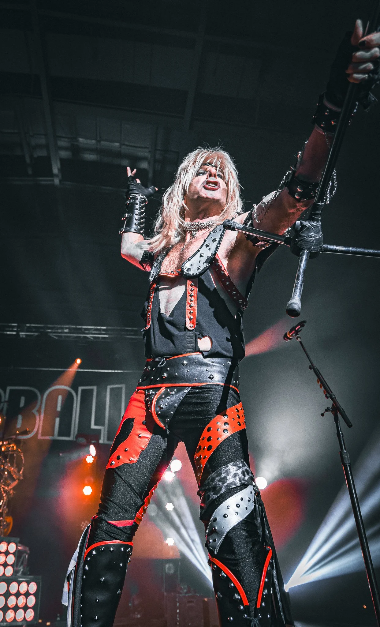 A performer on stage wearing black and red leather clothing with studs and chains, holding a microphone stand, with stage lights shining behind him, and a dark background.