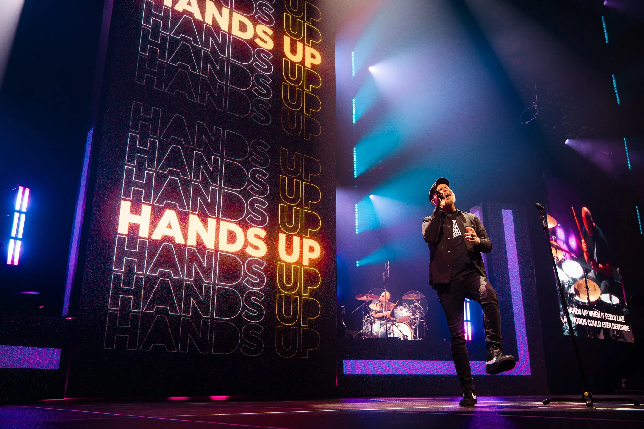 A singer performs on stage with a microphone, with colorful stage lights and a large screen displaying the words 'HANDS UP' repeatedly in bright and dark colors in the background. There are drums and additional instruments visible behind the singer.