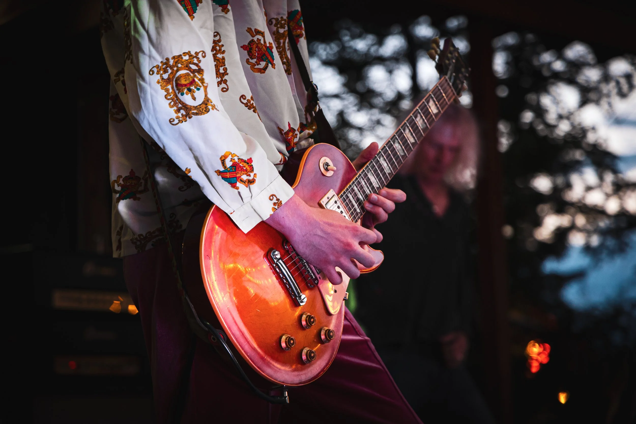 A musician playing an orange electric guitar at an outdoor concert during dusk, with another person blurred in the background.