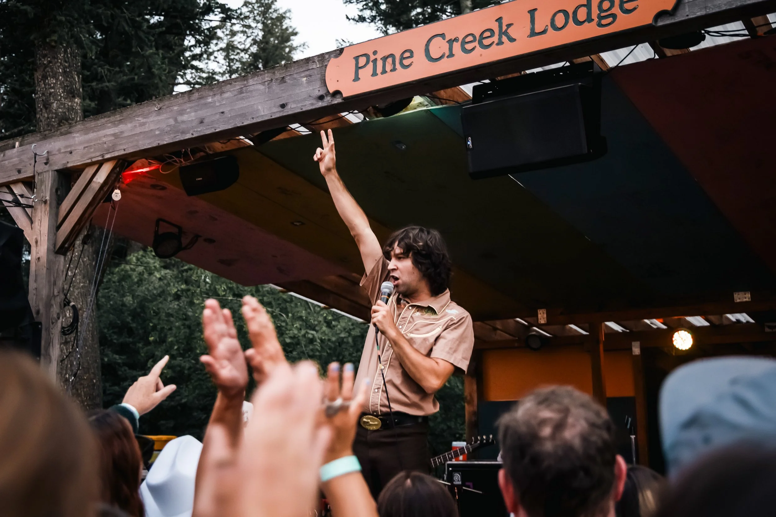 A man with dark curly hair, wearing a beige shirt, stands on a stage with a microphone in hand, raising his right hand with two fingers up, while performing in front of a crowd. The stage has a wooden structure with a sign that reads 'Pine Creek Lodge', and there's an outdoor setting with trees in the background.