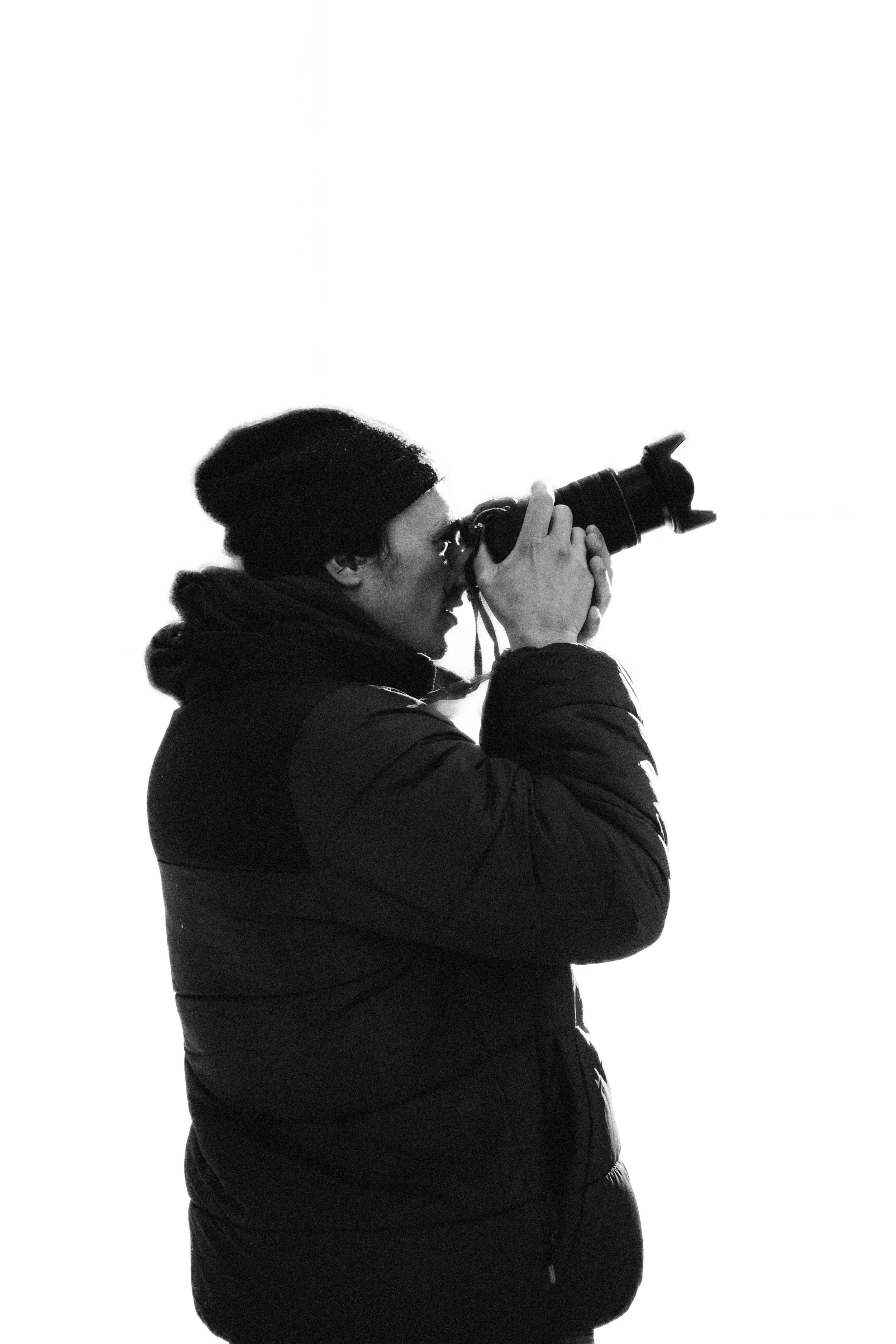 A person dressed in a winter jacket and beanie taking a photograph with a camera against a plain white background.