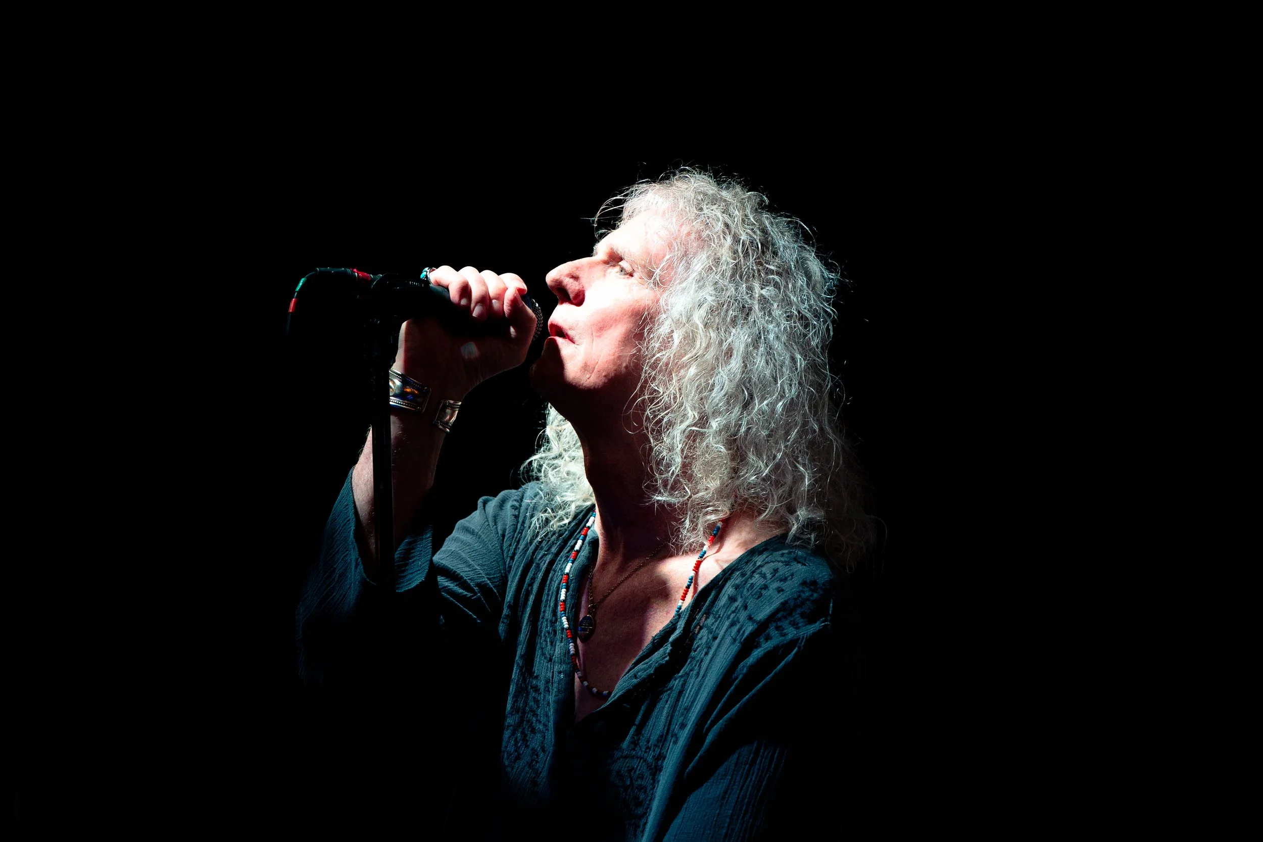 A woman with curly gray hair passionately singing into a microphone against a black background.