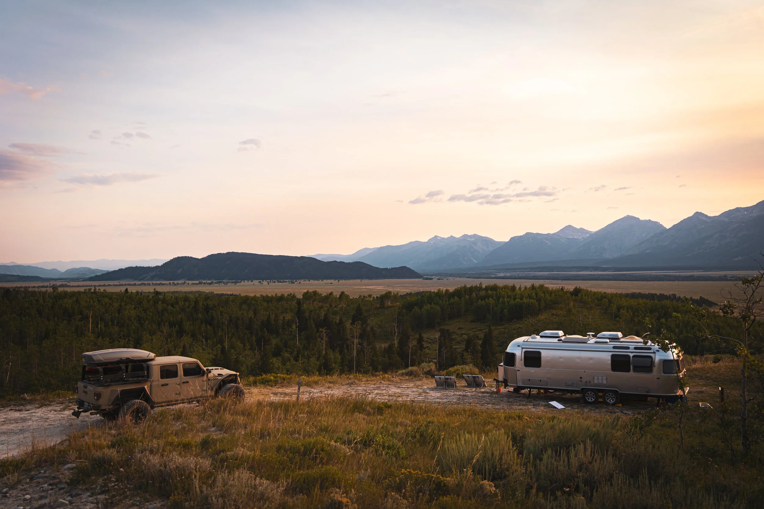A recreational vehicle and an off-road vehicle parked on a dirt area in a scenic landscape with mountains in the background during sunset.