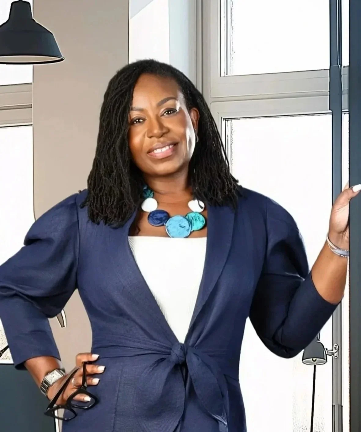 A woman with shoulder-length curly black hair wearing a navy blue blazer tied at the waist, colorful bead necklace, and silver watch, standing in front of a window, smiling at the camera.