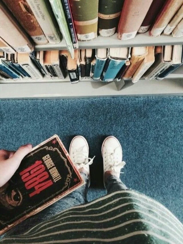 Instagram link; a person holds a copy of 1984 while standing in front of a shelf of library books.