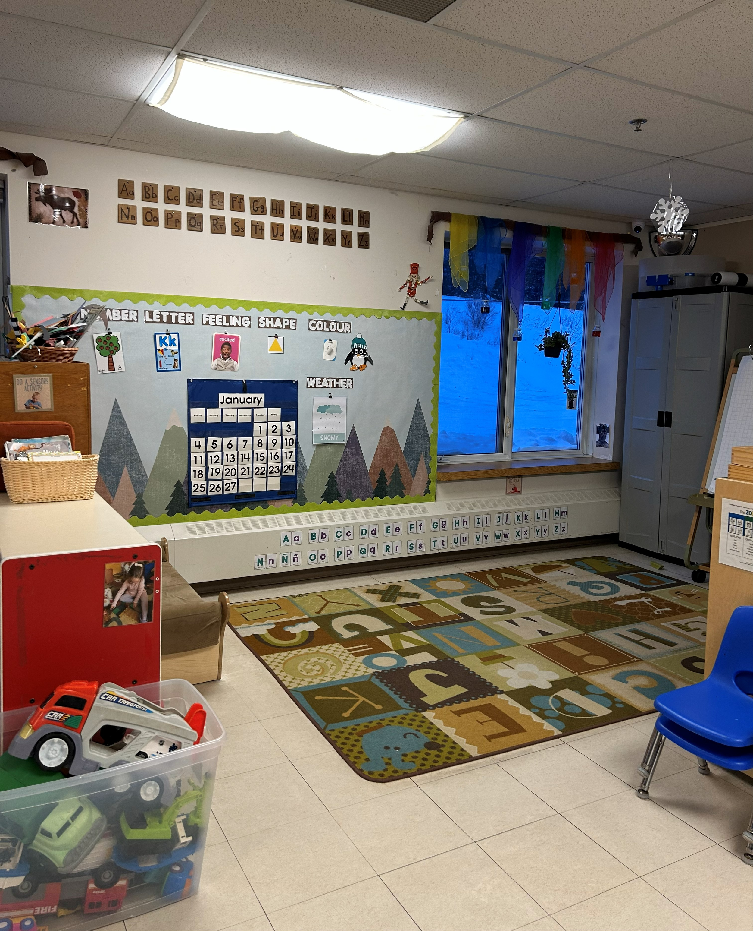 Classroom with colorful educational decorations, a calendar on the wall, and a carpet with alphabet letters. Toys are visible in a clear plastic container, and a blue chair is on the right side.