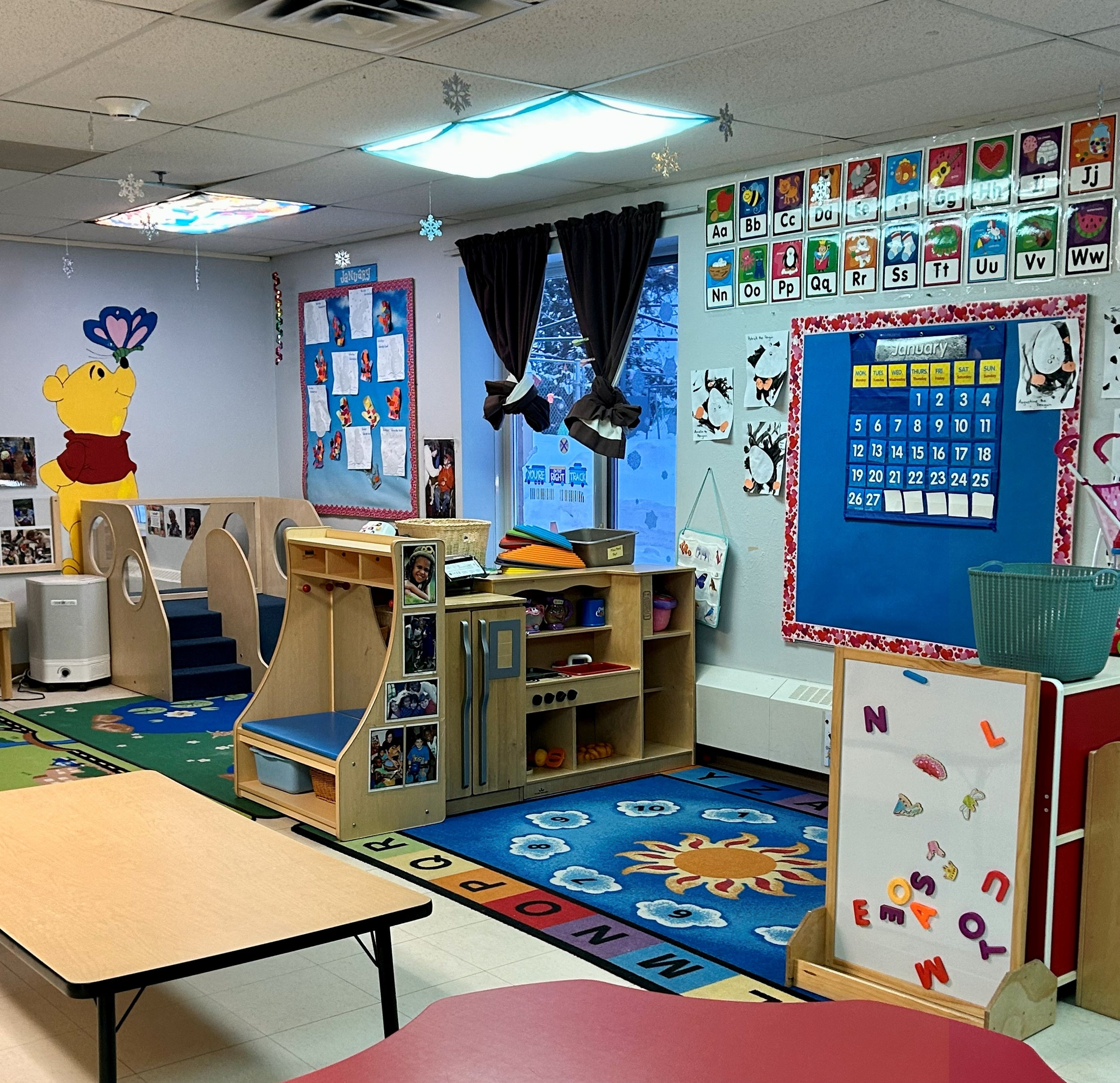 Colorful preschool classroom with wall decorations, a cartoon bear poster, a blue calendar, and a play corner with rugs and small furniture.