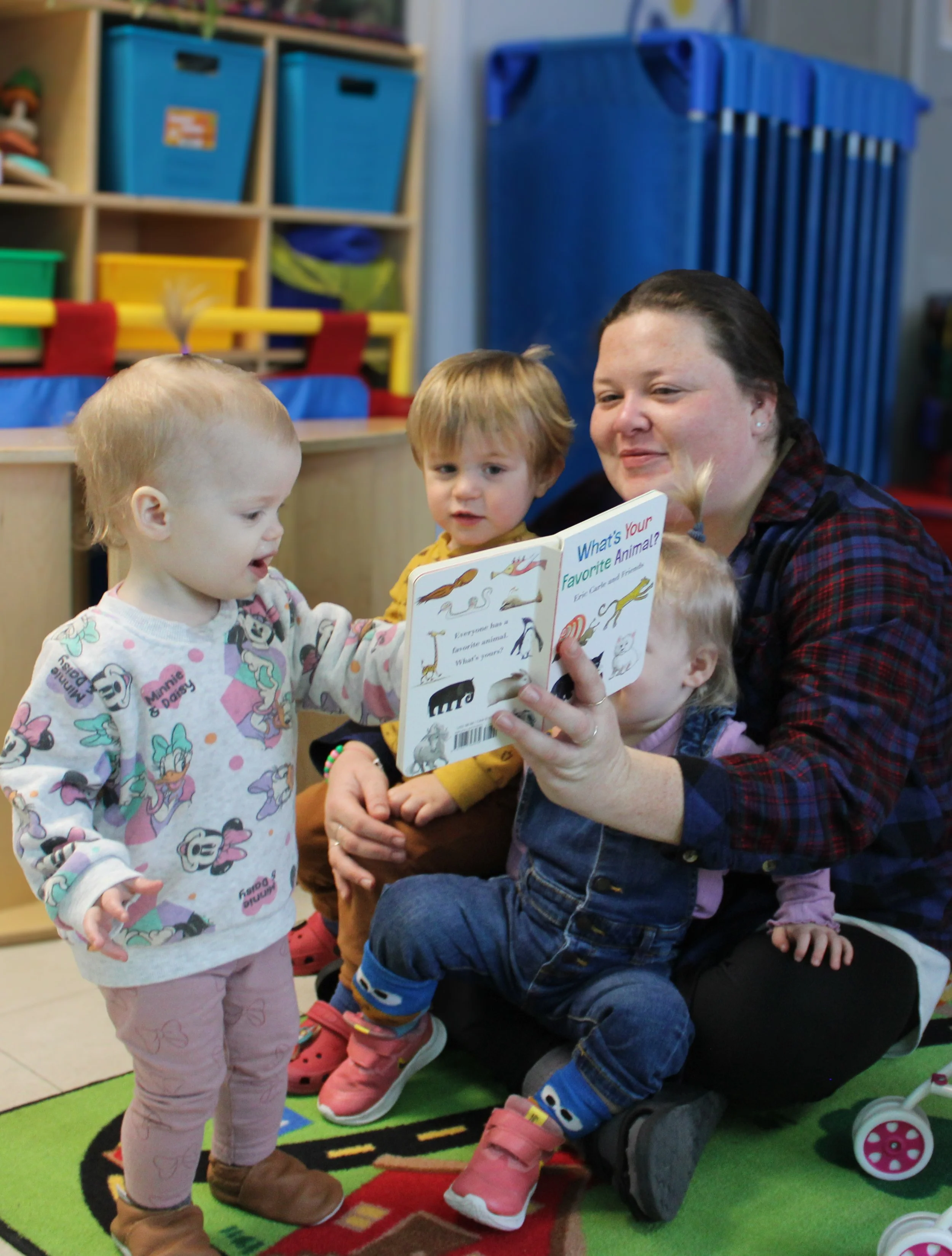 A woman reads a children's book titled "What's Your Favorite Animal?" to three young children in a playroom with colorful shelves and toys.