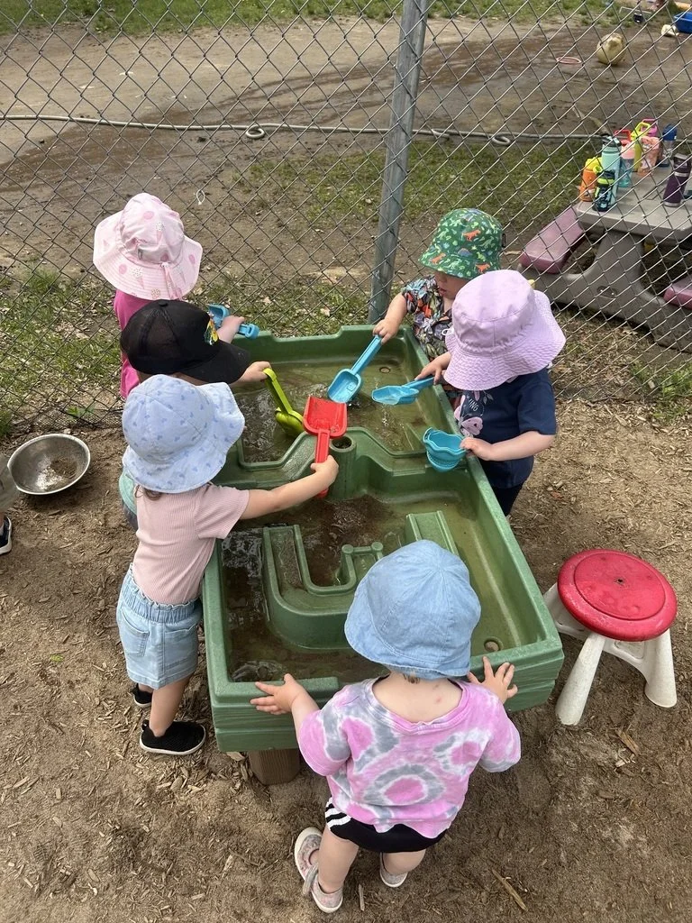 Six young children wearing hats and playing with sand and water in a sandbox outdoors, surrounded by a chain-link fence.