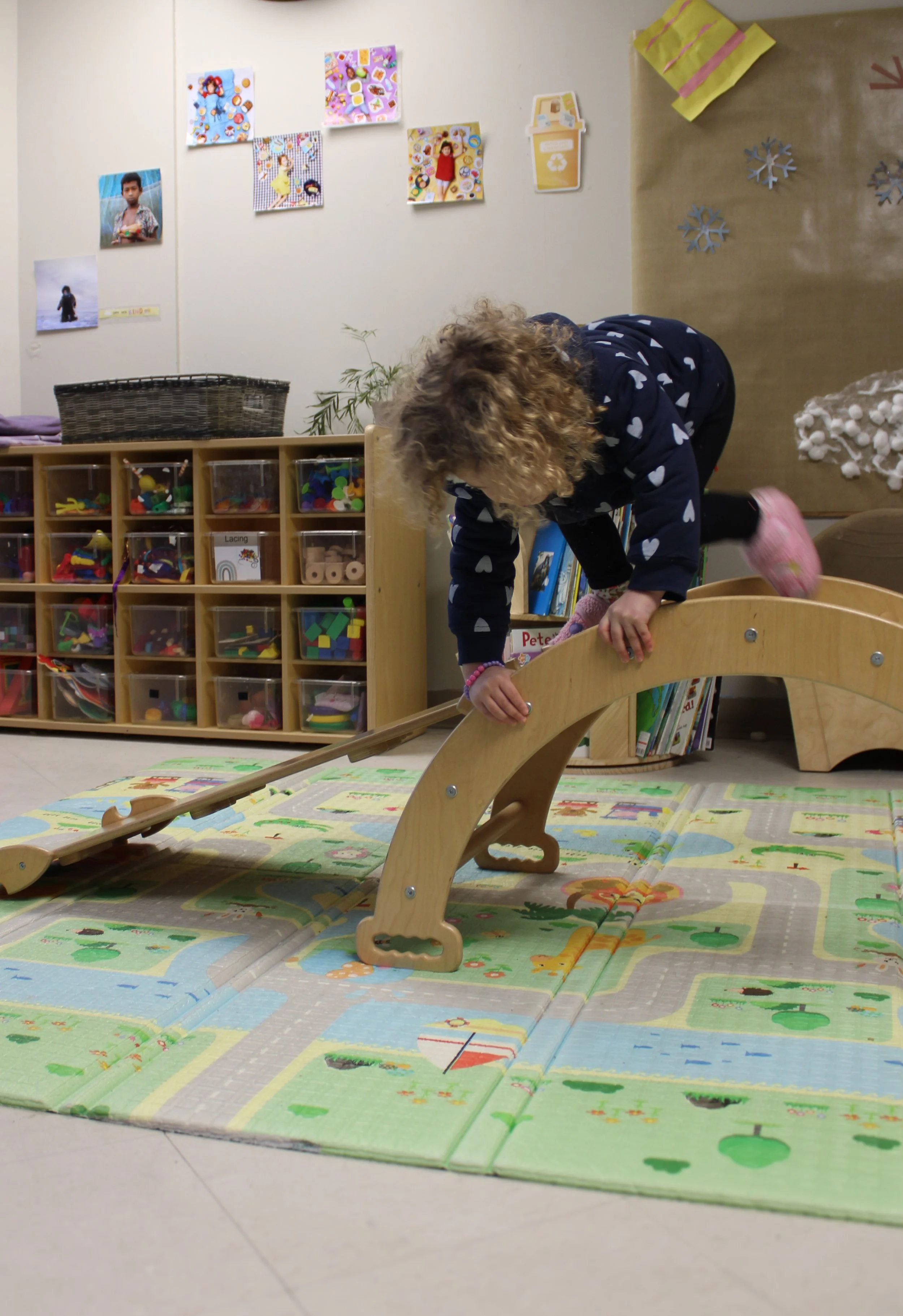 A young girl with curly hair is climbing over a wooden balance beam in a classroom or daycare setting.