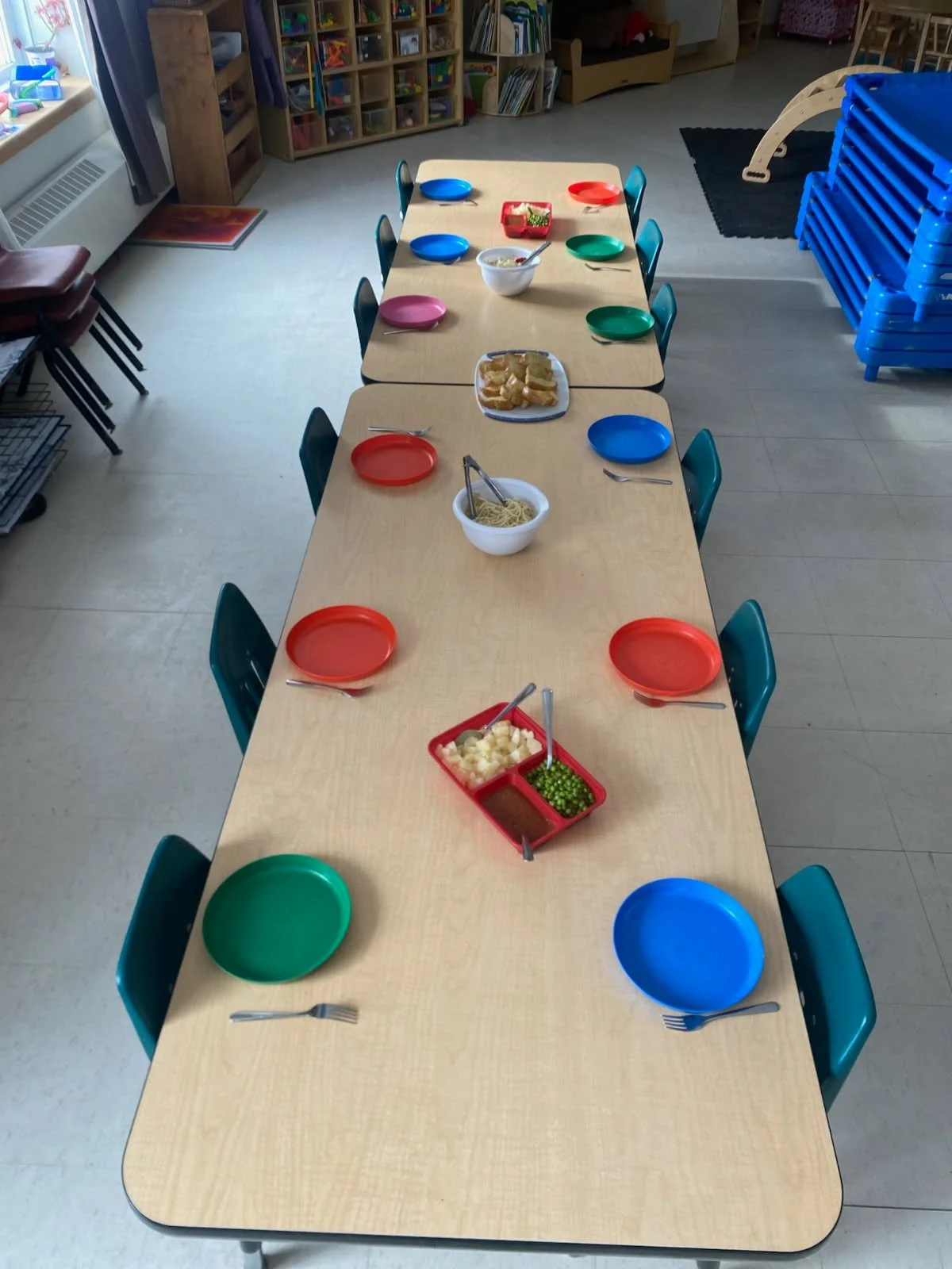 Children's table set for a meal with colorful plates and bowls, including salads, spaghetti, and bread, in a classroom or daycare setting.