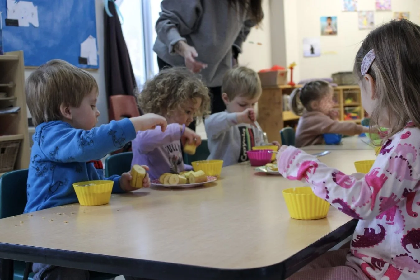 Young children sitting at a table in a classroom, eating sliced bananas from yellow bowls under the supervision of an adult.