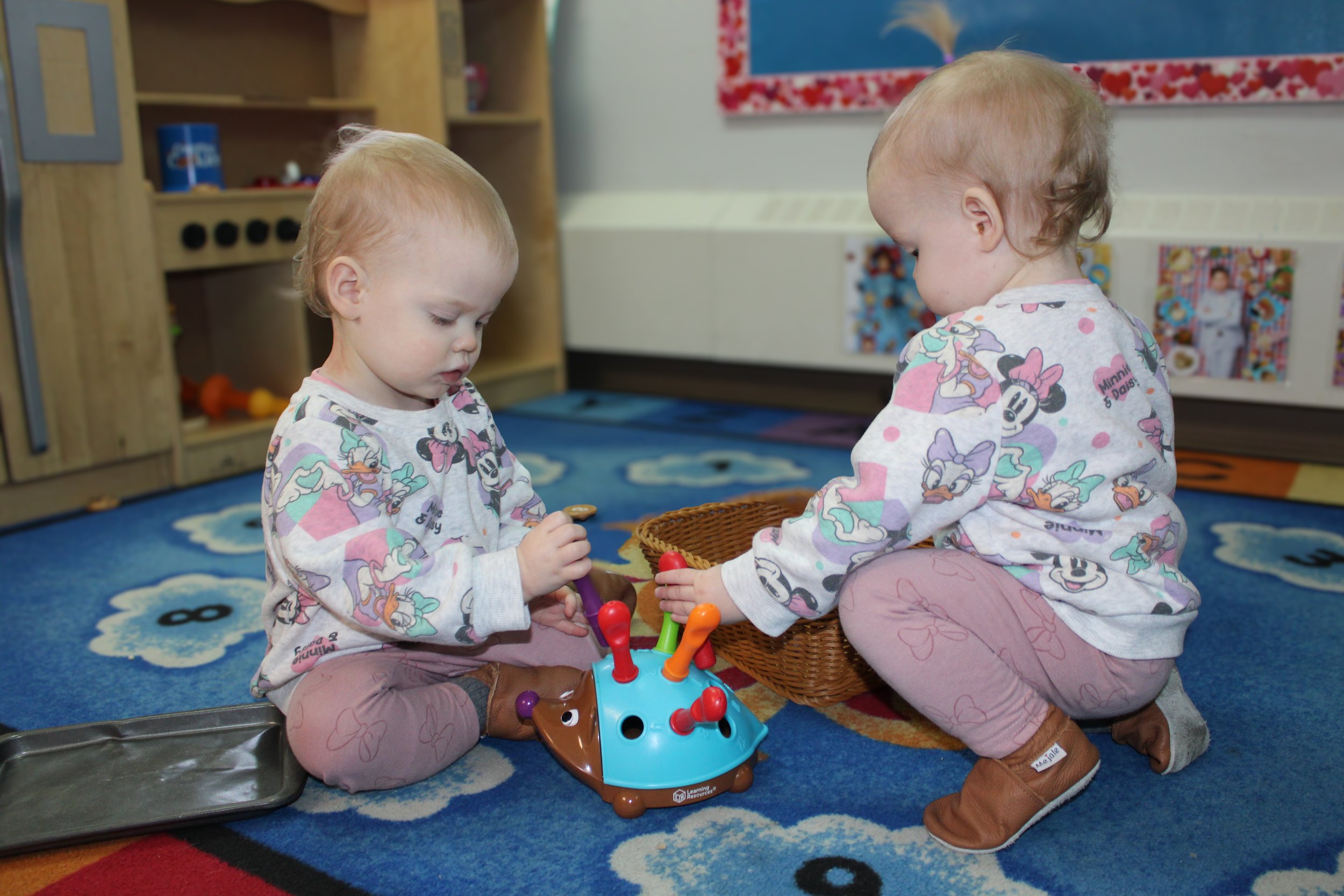 Two young children playing a game together on a blue educational rug in a classroom, with shelves and colorful decorations in the background.