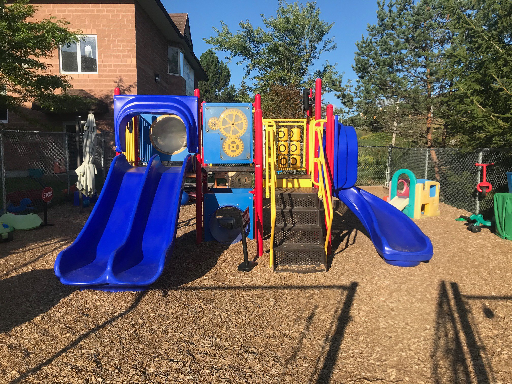Colorful playground with red, yellow, blue slides, climbing structures, and other play equipment on wood chip ground, fenced yard with trees and residential buildings in the background.