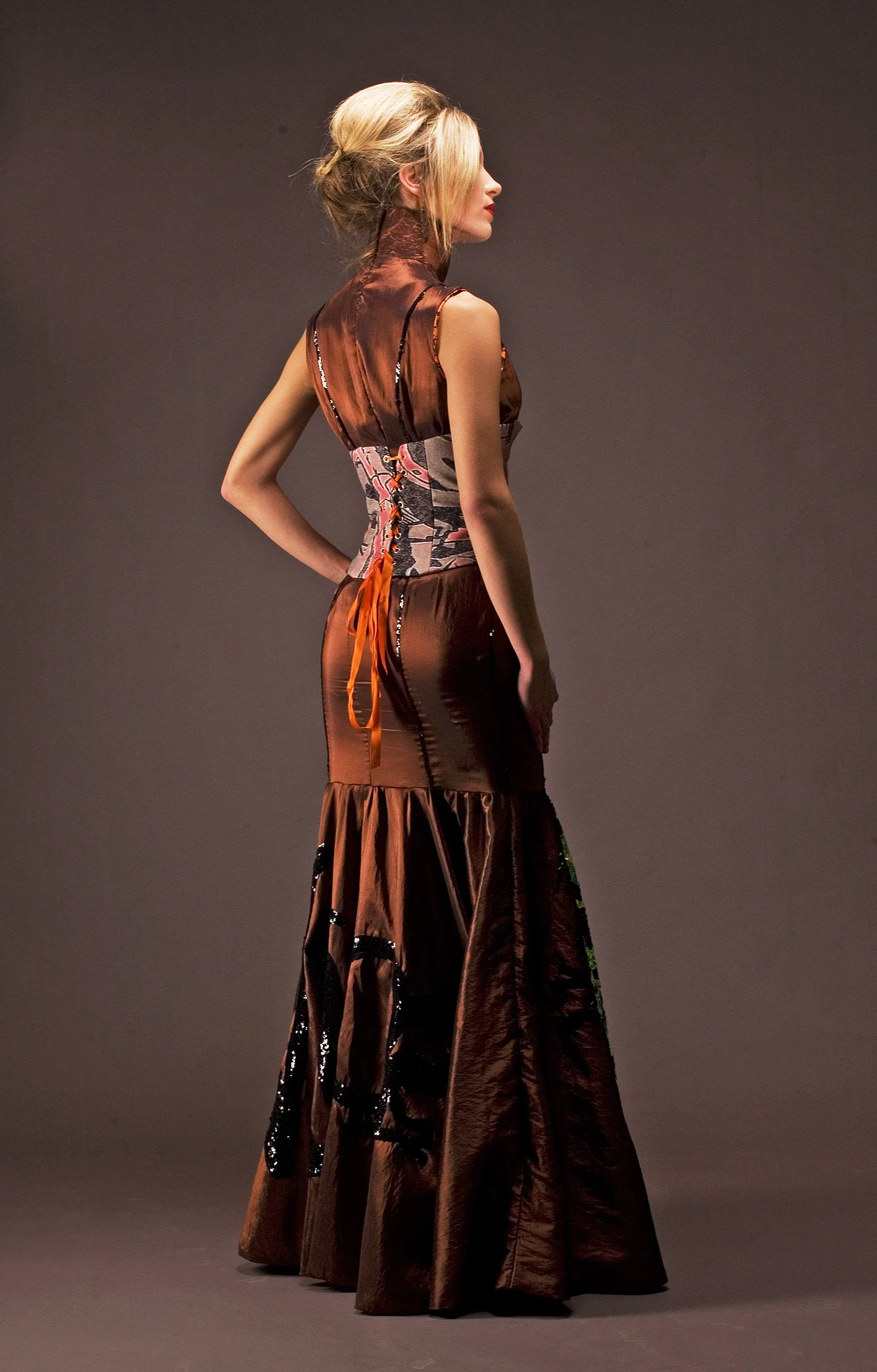 Side view of a woman with styled blonde hair wearing a sleeveless, brown satin dress with black and patterned details, standing against a plain dark background.