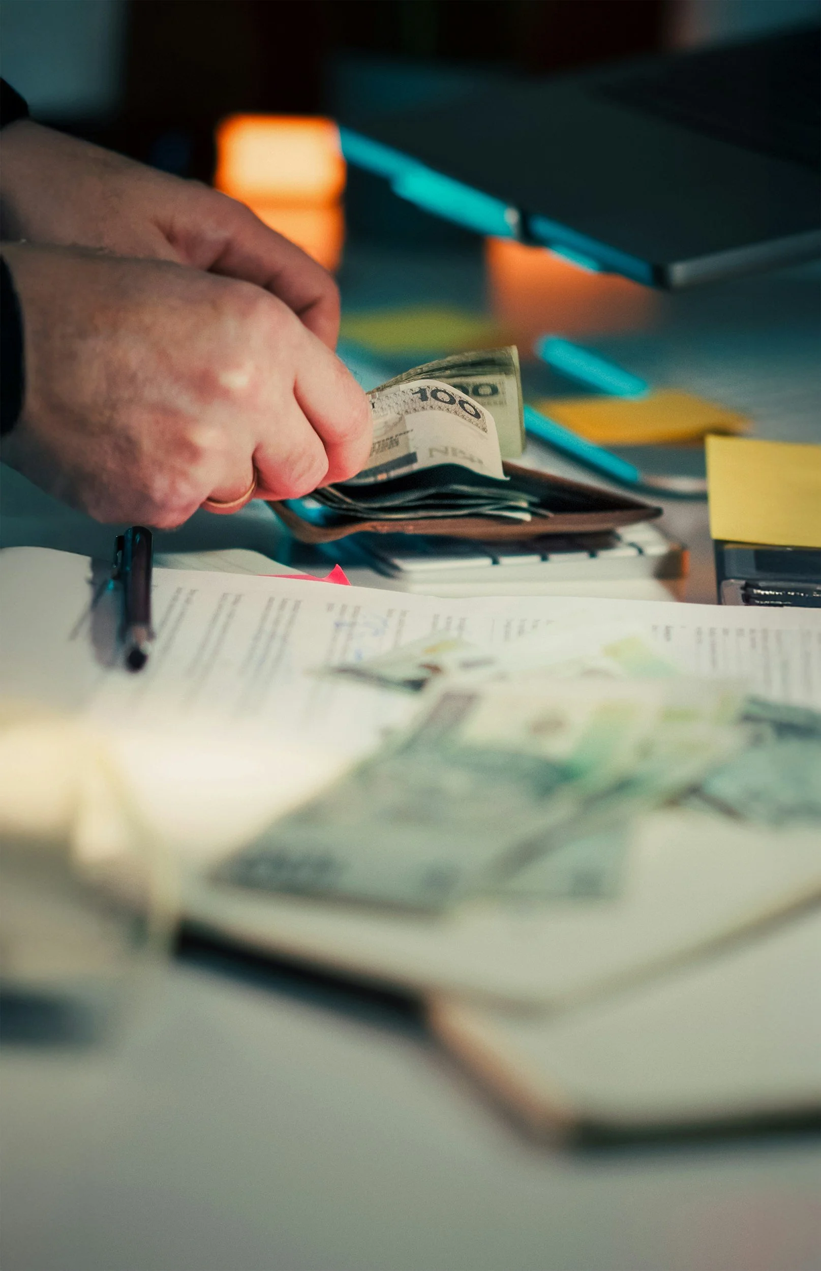 Close-up of hands counting dollar bills on a cluttered desk with sticky notes, pen, and open laptop.