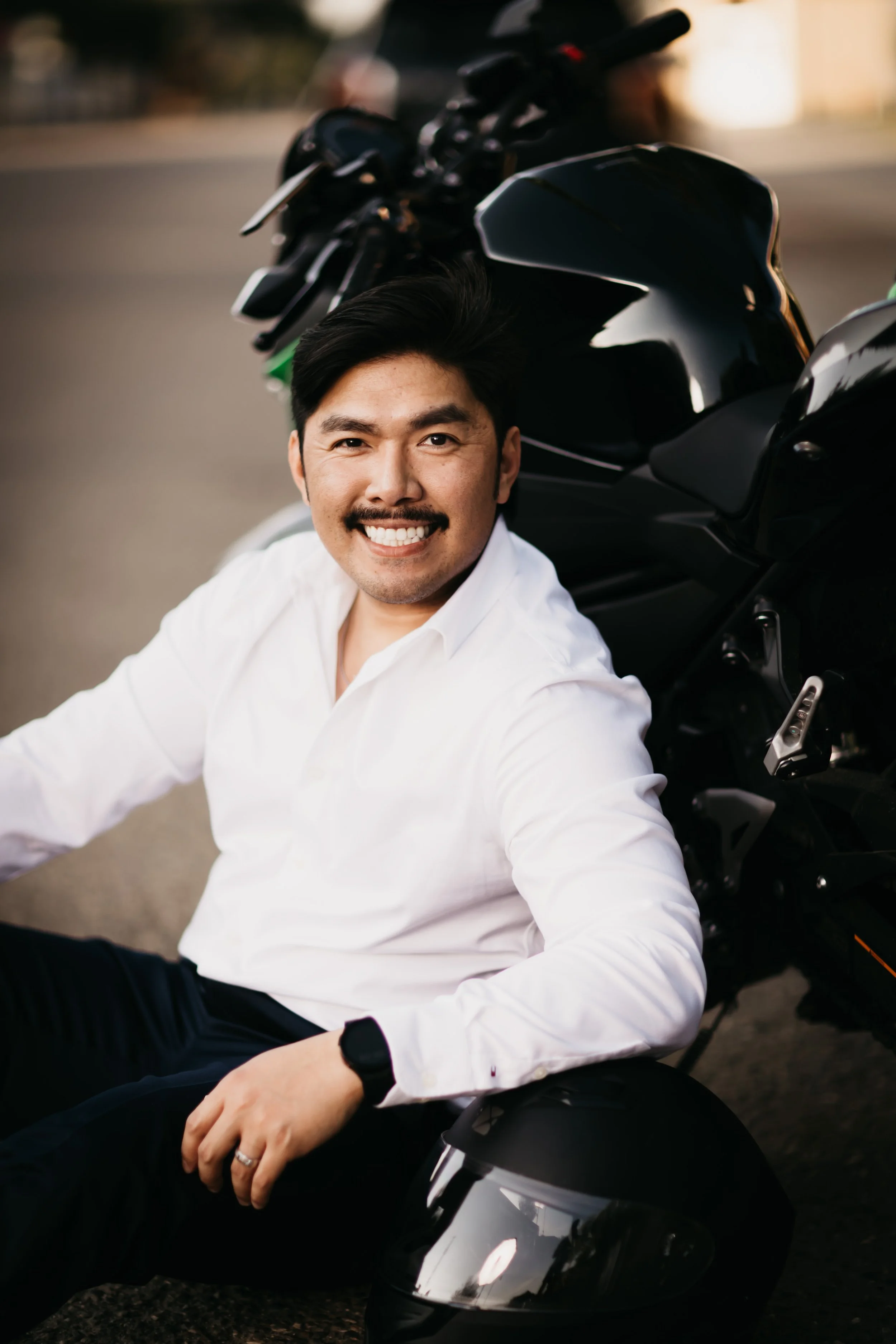 A man smiling and sitting next to a black motorcycle, wearing a white shirt and black pants.