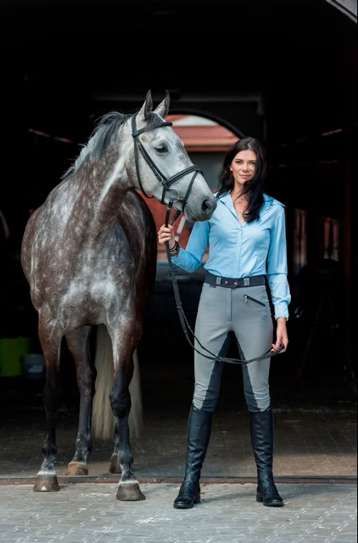 A woman standing beside a gray horse inside a stable, holding the horse's bridle.