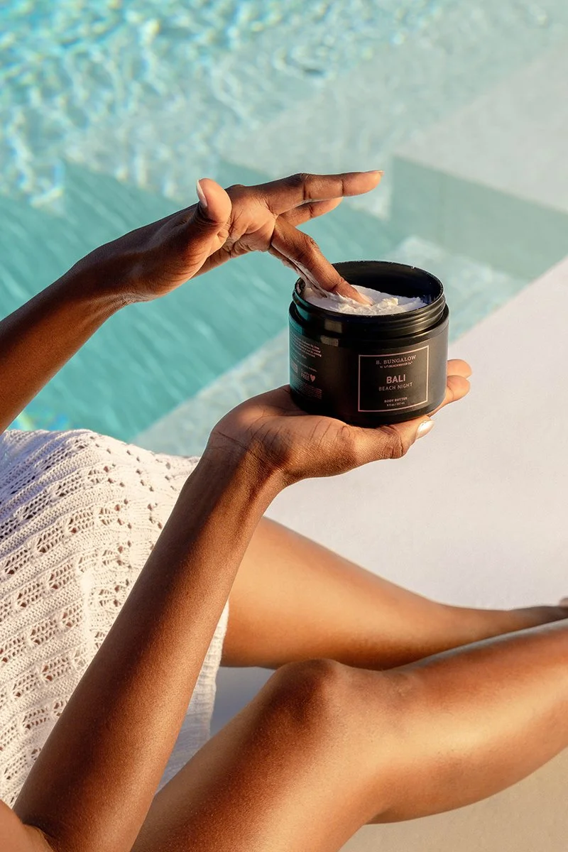 A person with dark skin is relaxing by a pool, applying body butter from a black jar labeled "Bali Beach Night" while sitting with their legs crossed and a white crocheted cover-up.