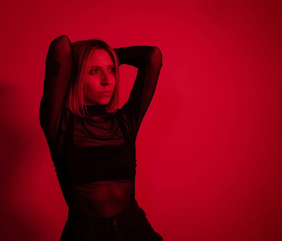 A woman with blonde hair wearing a sheer black top with long sleeves posing with her hands behind her head against a red background.