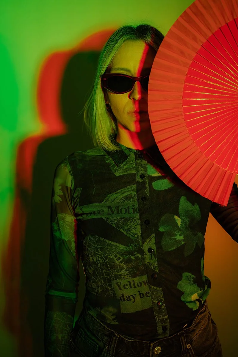 A woman with blonde hair wearing dark sunglasses and a patterned shirt, partially obscured by a large red paper fan, with colorful lighting casting shadows on her face and background.