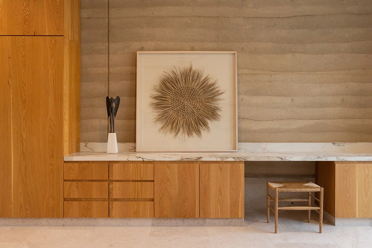 Interior view of a modern room featuring a wooden cabinet, a white marble countertop, a decorative sculpture placed in a white vase, a framed art piece with a sunburst design, and a small wooden stool.