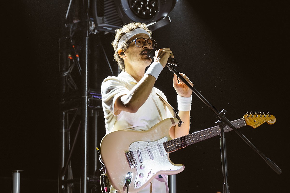 A musician wearing glasses, a headband, and white clothing playing an electric guitar and singing into a microphone on stage with lighting equipment behind them.
