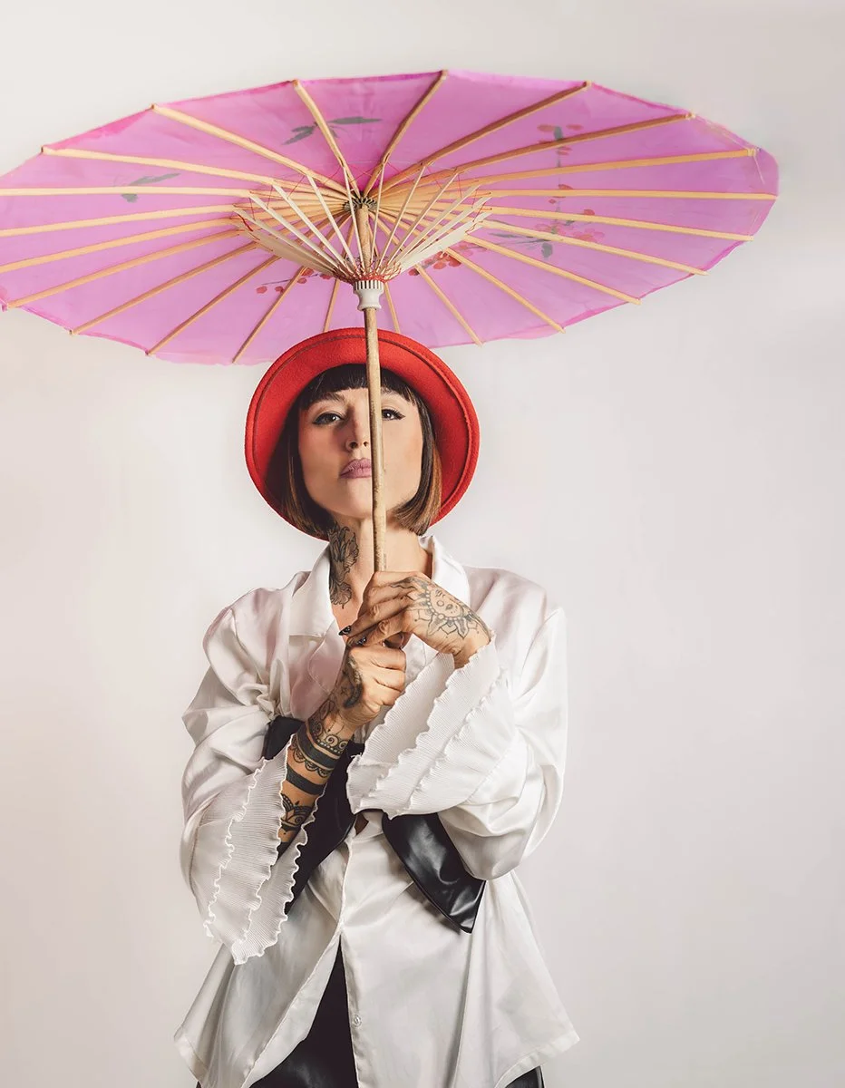 A woman with tattoos on her arms and neck, wearing a red hat and white shirt with ruffled sleeves, holding a pink parasol against a plain white background.
