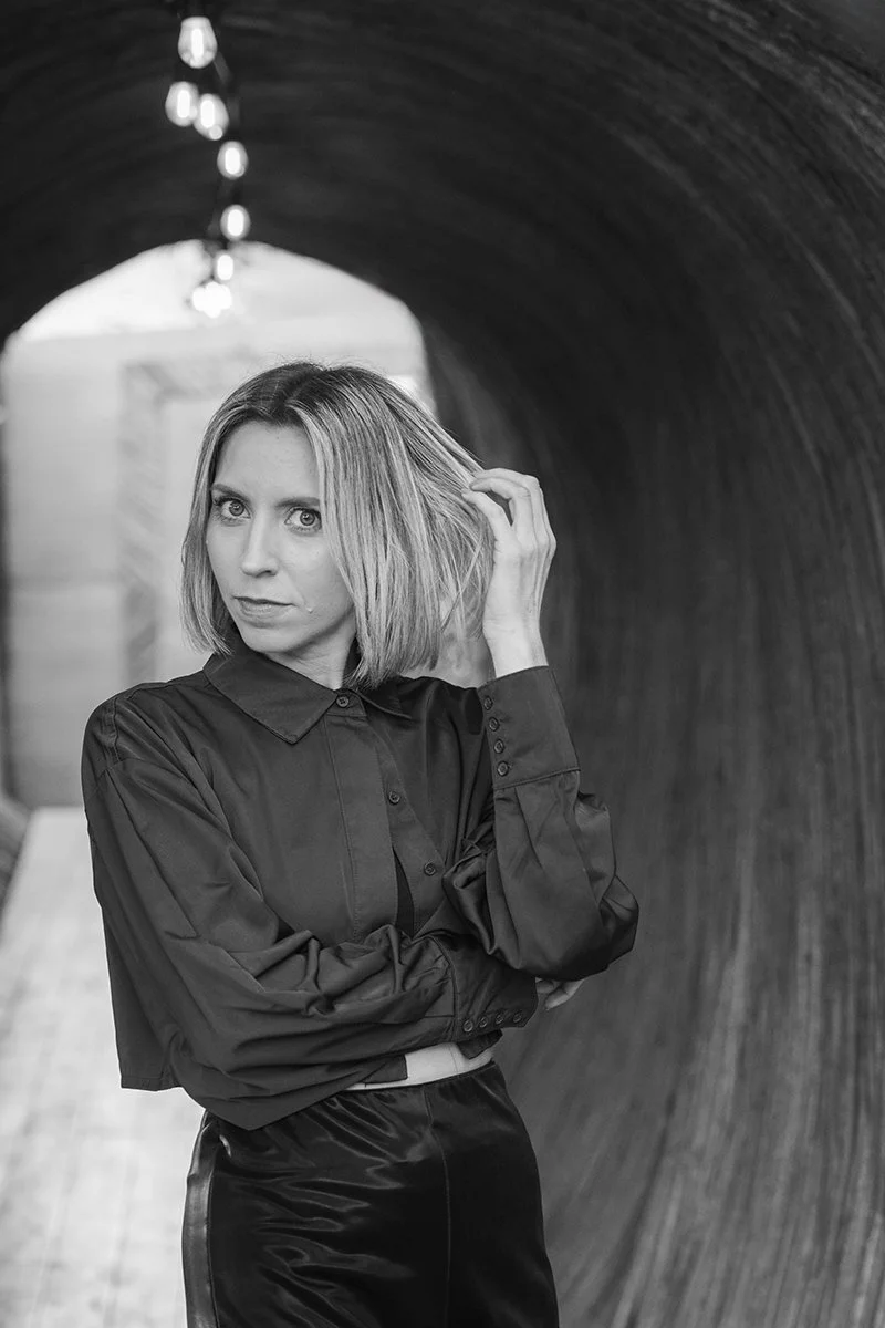 A woman with shoulder-length hair, wearing a dark button-up shirt, standing in a tunnel with lights hanging from the ceiling, looking at the camera and touching her hair.