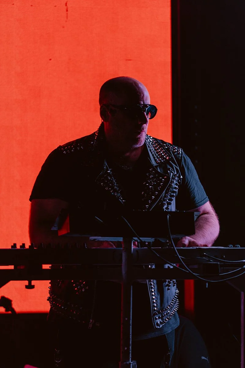 Bald man wearing sunglasses and a black leather vest with silver spikes, standing behind electronic music equipment, illuminated by red and orange background lights.