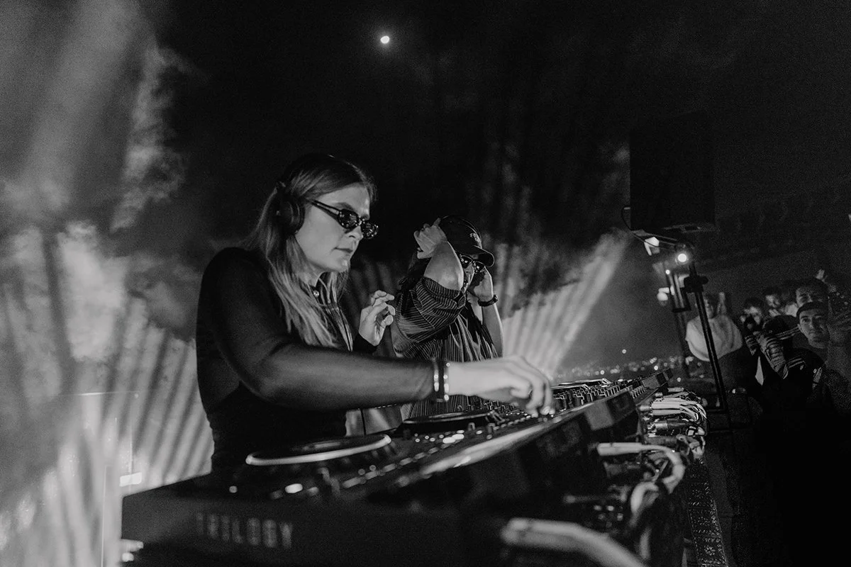 Two female DJs wearing headphones and sunglasses perform at a nightclub or concert venue, with an audience watching in the background, in black and white.