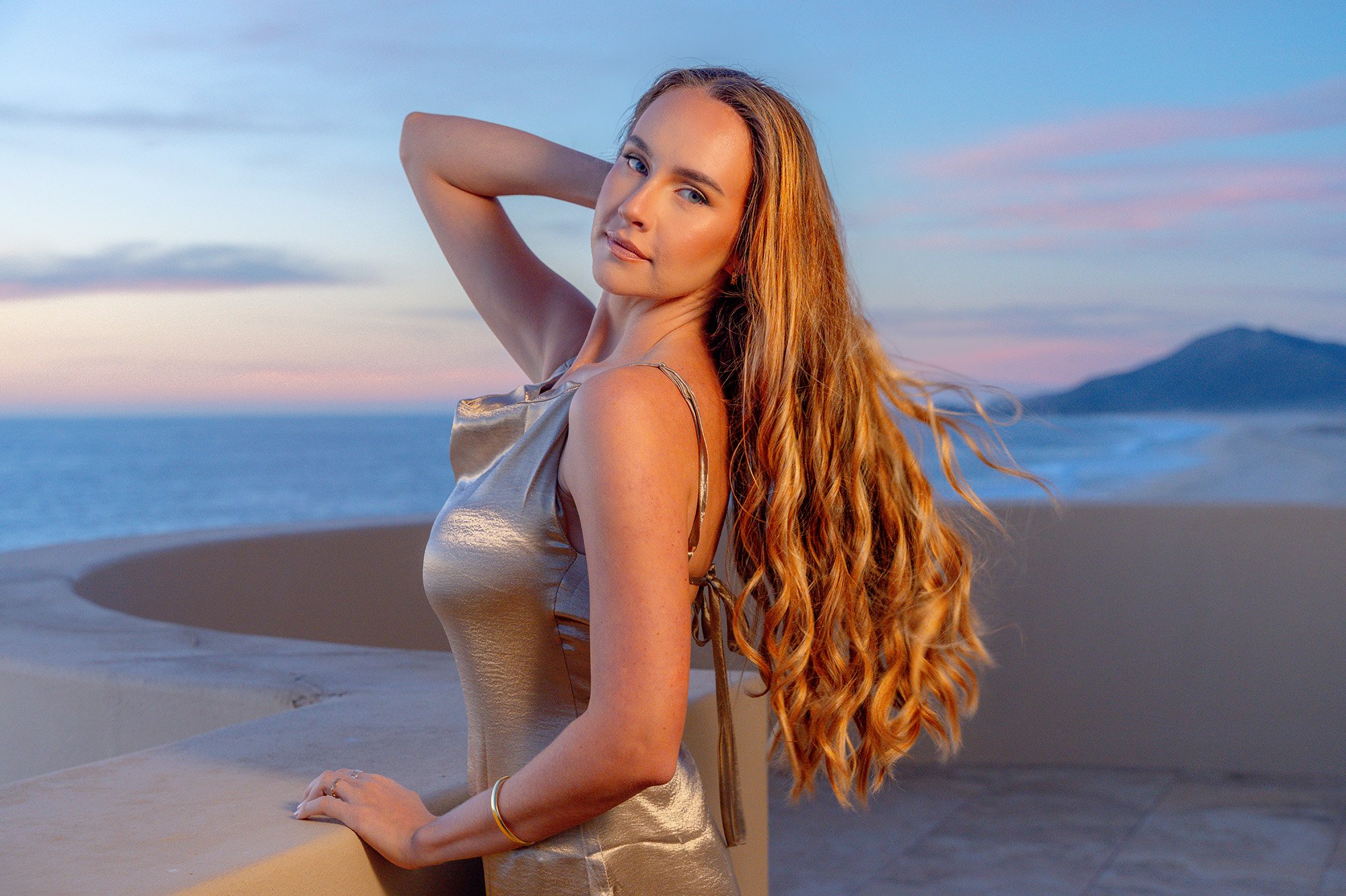 A woman with long red hair standing on a beach during sunset, looking at the camera with a serene expression.