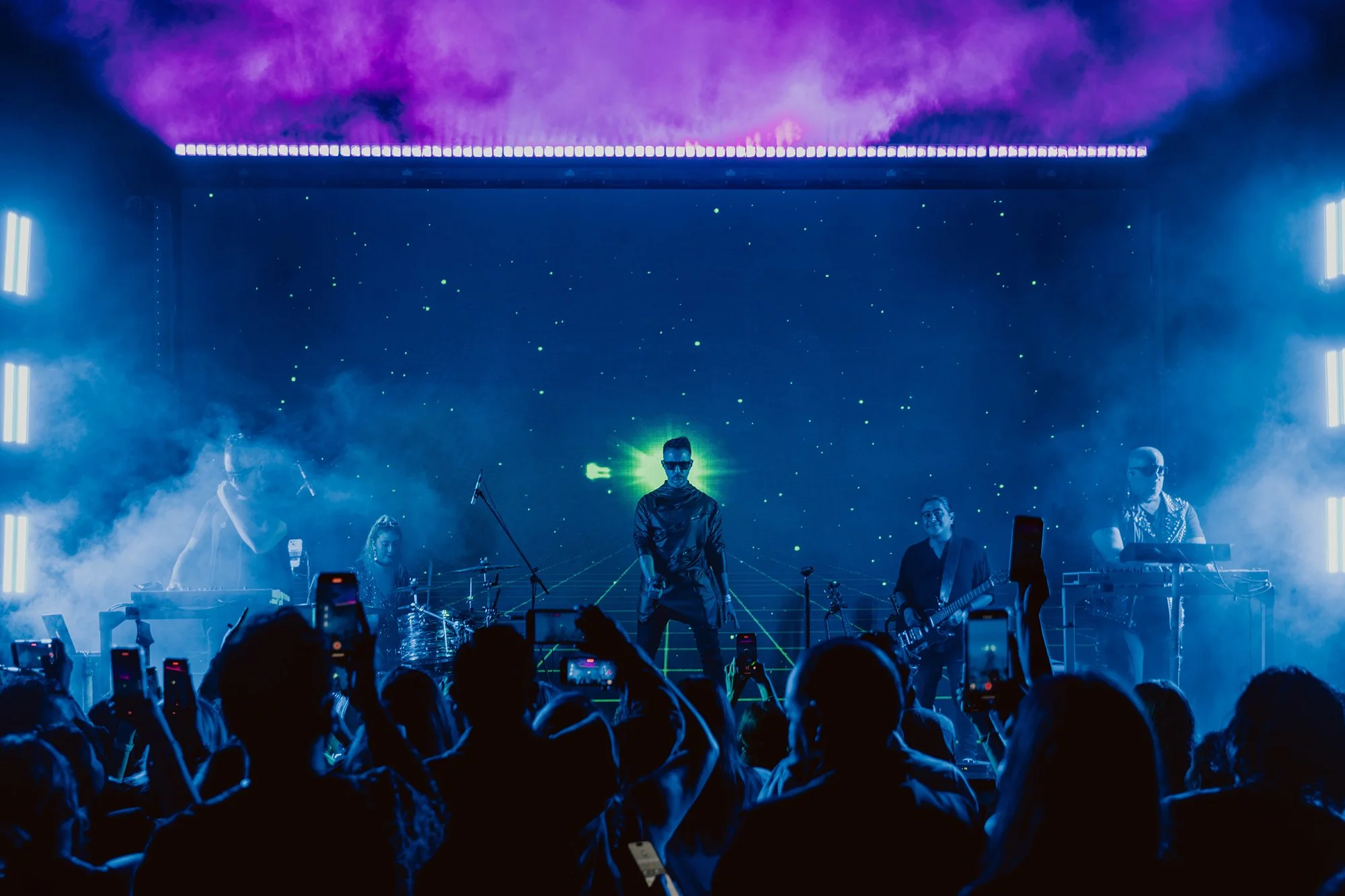 A live concert with a band performing on stage under vibrant purple and blue lights, surrounded by an audience capturing the moment with smartphones.