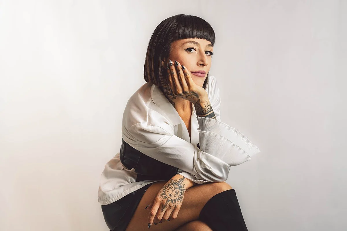 A woman with dark hair and bangs, tattoos on her hands and arm, sitting against a plain light-colored background, resting her chin on her hand.