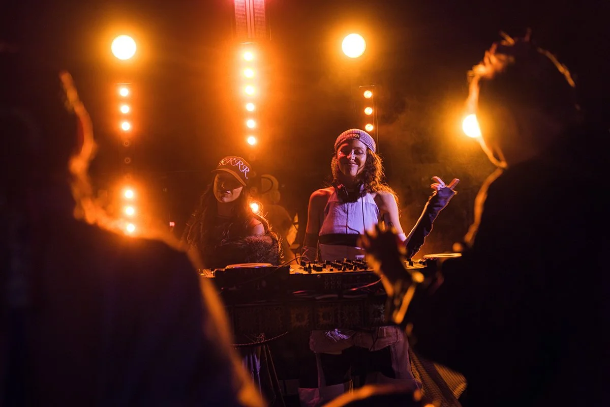 Women DJing at night with orange stage lights shining behind them.
