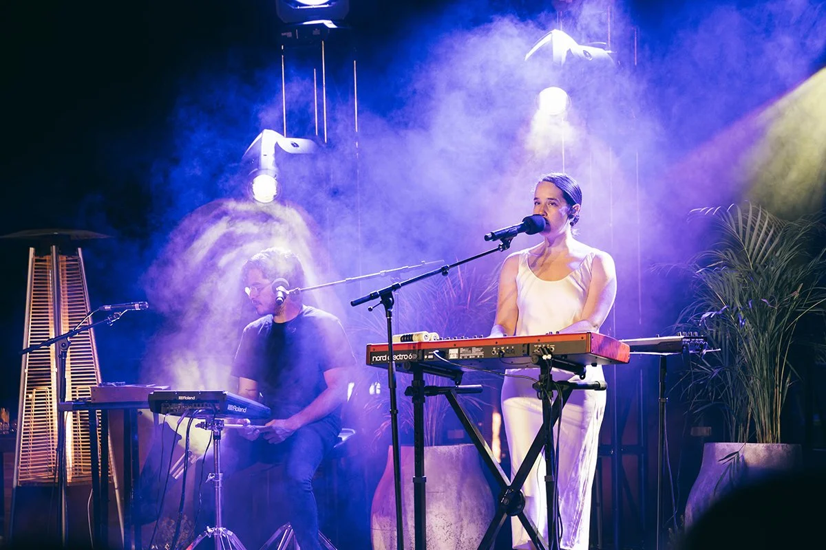 A woman and a man perform on stage with keyboards and microphones, surrounded by purple and yellow stage lighting and smoke.