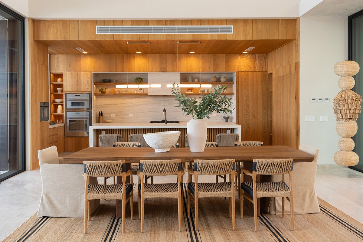 Modern dining area with a wooden table, wicker chairs, and a minimalist kitchen in the background featuring open shelves, built-in appliances, and a marble backsplash. A large white vase with green plants is on the table.