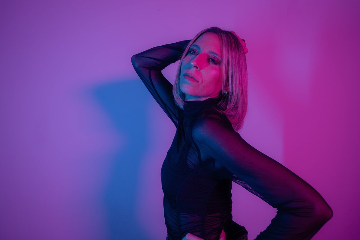 A woman posing against a purple and pink gradient wall, wearing a sheer black top with long sleeves and striking a confident pose.