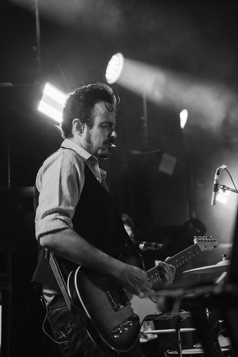 Man playing electric guitar on stage with stage lights and microphone in background.