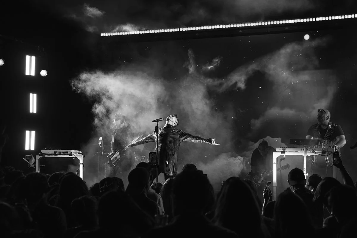 A musical live performance on stage in black and white, with a singer in the center performing with arms outstretched and a band playing behind them, audience members visible in the foreground.