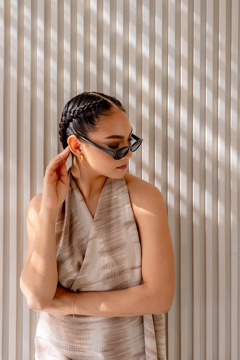 A woman with braided hair wearing sunglasses, gold earrings, and a sleeveless beige top, standing against a background of vertical white and beige striped wall.