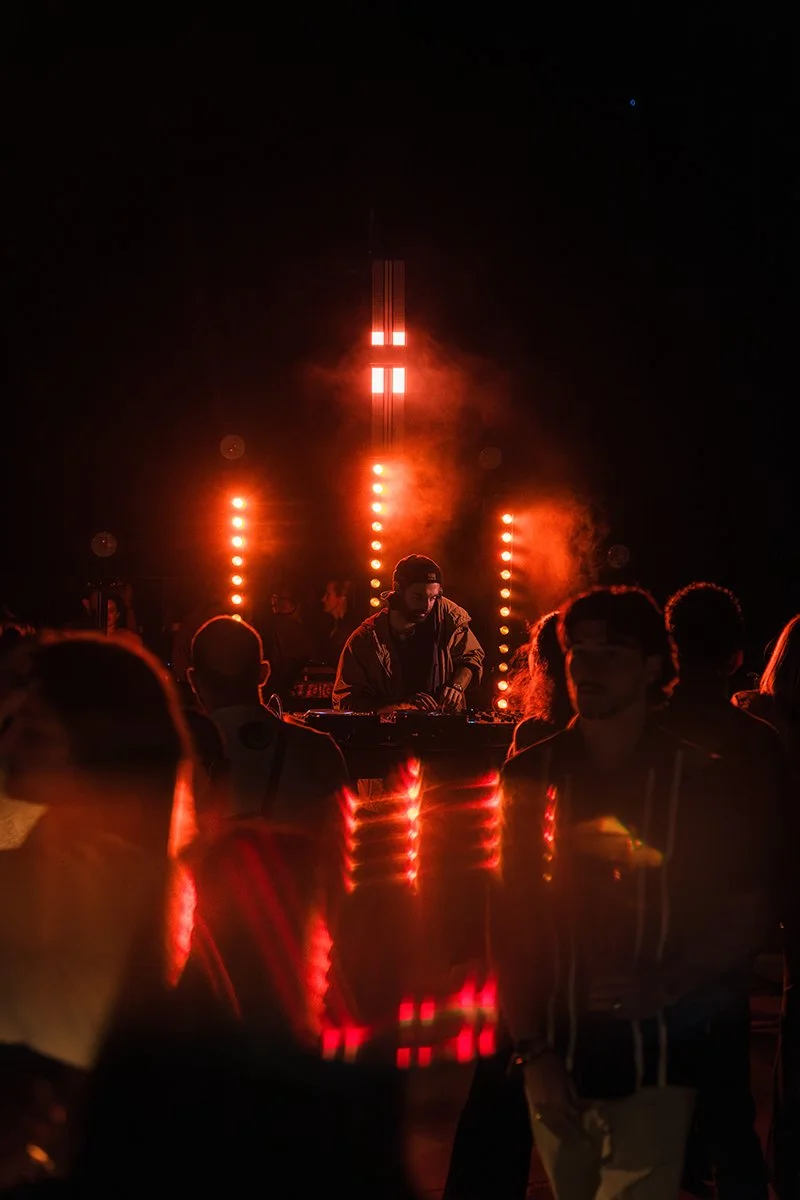 A DJ performing at a dark outdoor event with red and orange stage lights, smoke, and a crowd watching.