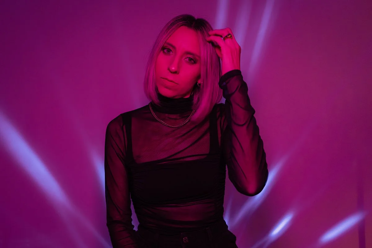 Woman with shoulder-length blonde hair wearing a sheer black top, standing against a purple and pink lighting background.