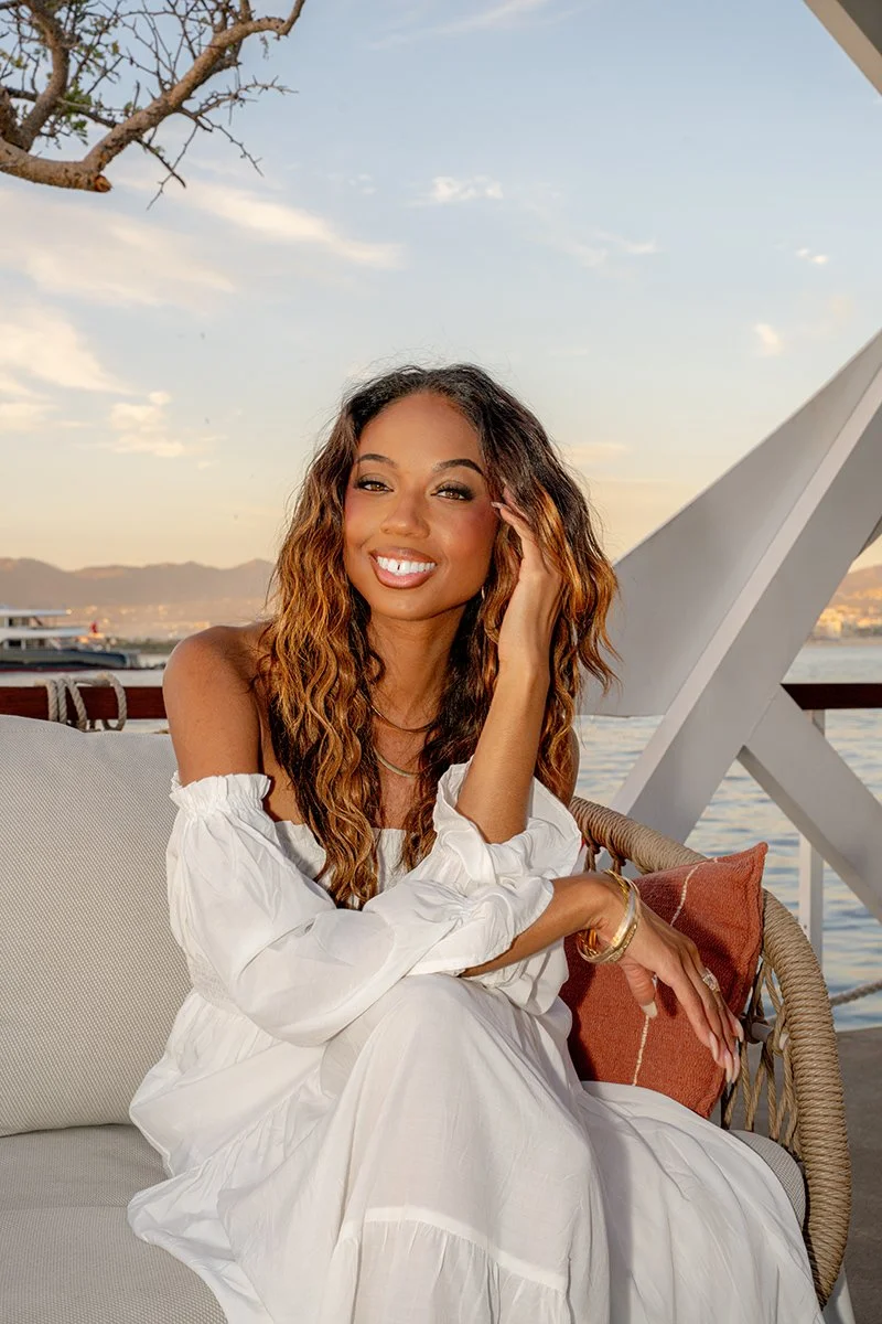A woman with long wavy hair and a white off-the-shoulder dress, sitting outdoors on a boat deck with water and city skyline in the background, smiling at the camera.