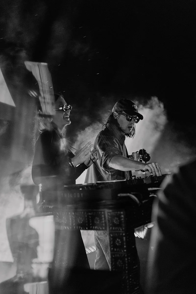 Black and white photo of a DJ mixing music at a nightclub or concert, with a woman dancing and smiling in the background, both wearing sunglasses.