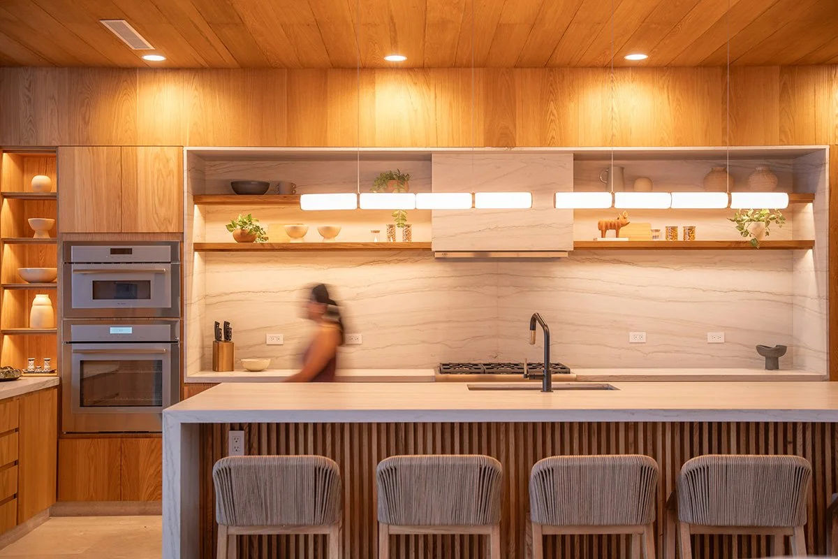 Modern kitchen with wooden cabinetry, marble backsplash, and open shelves displaying bowls, plants, and decor. An island with four chairs and a sink in the foreground. Blurred person walking in the background.